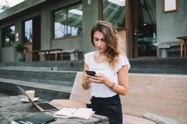 Focused busy adult woman standing beside table with laptop and open notebook outside of single-storey building and texting on smartphone