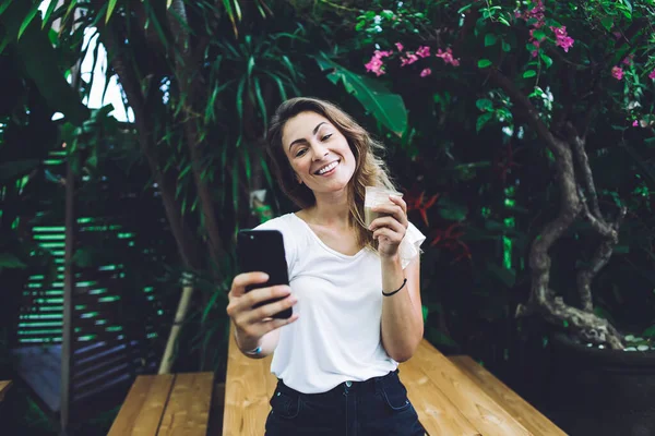 庭の木のテーブルの近くに立っている白いTシャツに肯定的な女性を笑顔とスマートフォンで飲むのガラスで自撮り — ストック写真