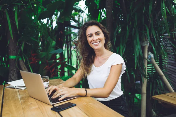 Joyful content woman relaxing in open air restaurant while sitting at table with smartphone and notebook and using laptop and looking at camera