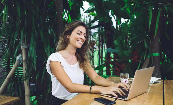 在热带咖啡馆的阳台上 坐在桌子旁 微笑着看着笔记本电脑屏幕 在键盘上打字的快乐而迷人的自由职业女性 — 图库照片