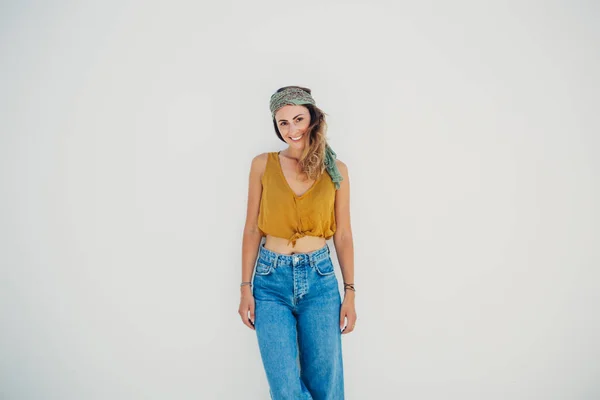 Open minded woman with charming smile in casual stylish wear standing with crossed legs along grey wall and looking at camera