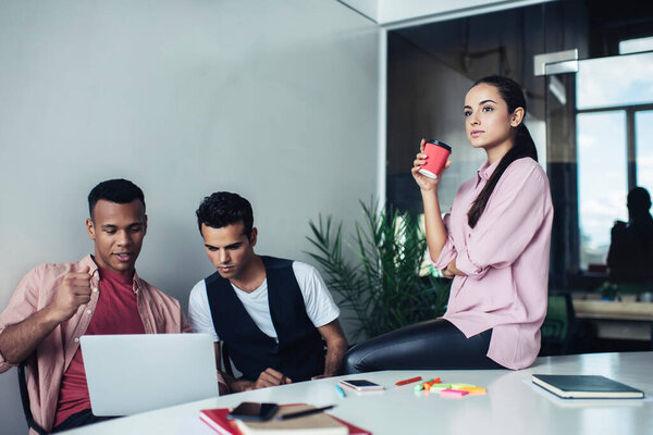 Многорасовая интеллектуальная группа сотрудников в повседневной одежде, сидящих за столом со смартфоном и чашкой кофе в современном светлом креативном офисе 