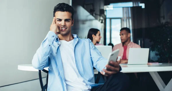 Portrait of happy millennial man holding cellphone device for communication in hand and smiling at camera during work day in office space, Spanish generation Y enjoying job experience indoors