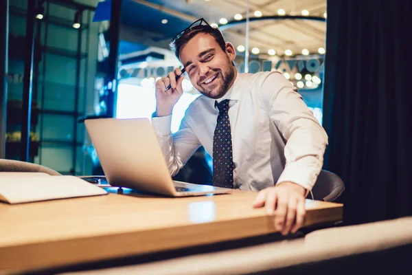 在现代有创意的办公室工作场所 穿着白衬衫 打着领带 头戴笔记本电脑 头戴书本的快乐男人坐在木制桌旁 — 图库照片