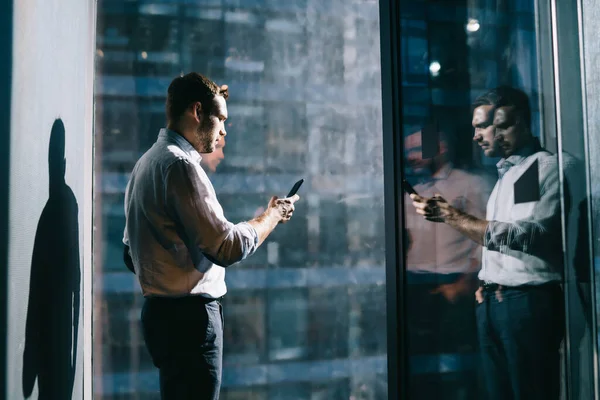 オフィス空間のガラスを反射する大きな窓の近くに立つスマートフォン上の正式なシャツのメッセージで若い深刻な男性に集中 — ストック写真