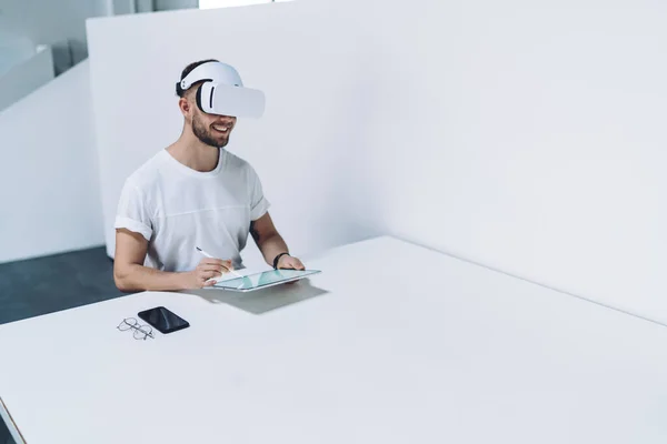 穿着白衬衫 面带微笑的年轻人坐在桌旁 戴着虚拟现实的耳机 一边用钢笔在平板电脑上画画 — 图库照片