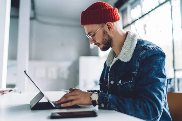 Tarz sahibi, sakallı, gözlüklü ve şapka takan genç bir erkek öğrencinin beyaz odadaki tablete mesaj yazışının yan görüntüsü bulanık arka planda.