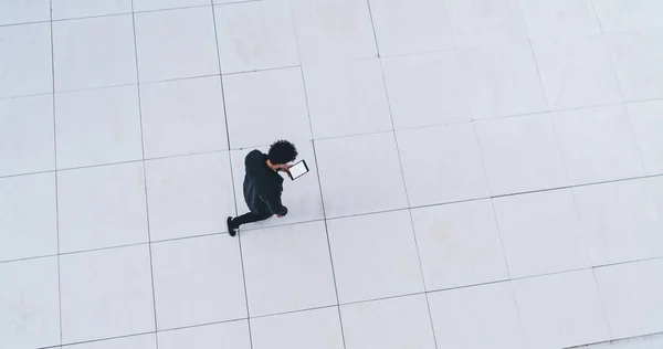 Top view of faceless man in black clothes walking on checkered floor near white wall and texting on tablet with blank space