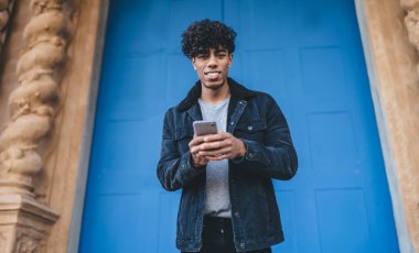 Siyah ceketli, kot pantolonlu, gülümseyen, kıvırcık, etnik bir adam sokak mavi kapısının önünde kameraya bakarken akıllı bir şekilde sörf yapıyor.