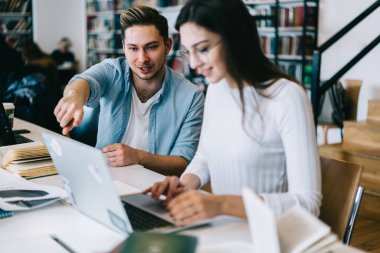 Hevesli öğrenci dizüstü bilgisayarında hata yapıyor ve kütüphanede açık kitaplarla dolu bir masaya parmağıyla işaret ediyor. 
