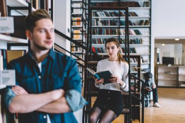 Entellektüel zeki, gündelik giysiler içindeki genç öğrenciler büyük şehir kütüphanesinde kitaplıkların karşısında duran kitapları seçip okuyorlar.