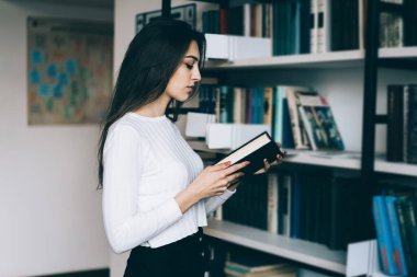Uzun siyah saçlı, beyaz kazaklı, akıllı genç bir kadın kütüphanede kitaplığın karşısında kitap okuyor.