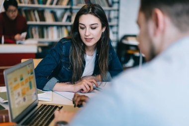 Kütüphanede okurken laptopta çok ırklı öğrencilerin bilgi daktilolarını izleyen zeki, uzun saçlı bir öğrenci. 