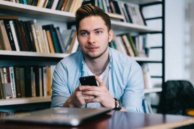 Kot gömlekli, beyaz tişörtlü gülümseyen genç adam kütüphanede cep telefonuyla mesajlaşıyordu.