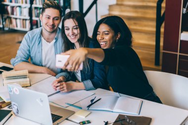 Kütüphanede kitaplarla ve laptopla çalışırken ve çalışırken cep telefonunda grupla selfie çeken neşeli, çoklu etnik kökenli uzun saçlı kadın. 