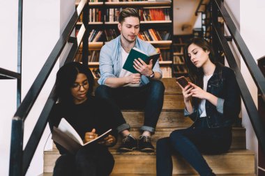 Aşağıdan gelen konsantre genç kız öğrenci akıllı telefon kullanırken, siyah kadın not yazıyor ve erkek kütüphanedeki merdivenlerde ders çalışırken kitap okuyor.