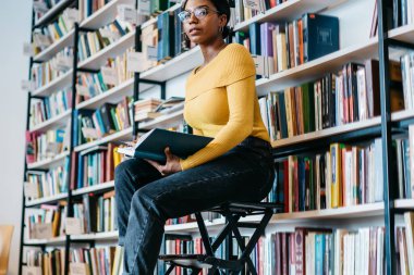 Üniversite kütüphanesindeki kitaplığın yanında, merdivende oturmuş ders kitabı okurken, günlük kıyafetli ve gözlüklü zeki siyahi bir kadın kameraya bakıyor.