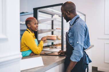Üniformalı Afro-Amerikan genç bir adamın pastaya bakıp sarı gömlekli ve turkuaz önlüklü şık siyahi bir kadınla konuşması. 