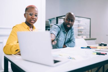 Ciddi Afro-Amerikalı bir adam, kadın meslektaşıyla birlikte kahve dükkanında iş projesinde çalışırken dizüstü bilgisayar monitörünü işaret ediyor.