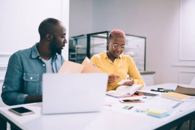 Düşünceli Afrikalı Amerikalı adam ekrana odaklanırken, bayan meslektaşı da akıllı telefon kullanarak modern kafede kağıt ve dizüstü bilgisayarla masada oturuyor.