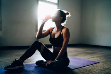 Arka ışıklandırma çekici kadın rahat spor giyimli rahat bir kadın yoga minderinde oturup gölgeli spor salonunda şişeden su içiyor.