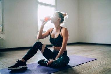 Spor giyim kuşağında arka ışıklandırmada çekici genç bir kadın yoga minderinde oturuyor ve gündüz vakti spor salonunda şişeden saf su içiyor.