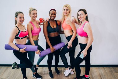 Modern spor kıyafetleri içindeki iyimser kadınlar spor salonlarında spor aksesuarlarıyla dikilip kameraya bakarken kucaklaşıyorlar.