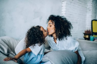 Evdeki kanepede dinlenirken küçük tatlı bir kızın etnik anneyi dudaklarından öpmesi.