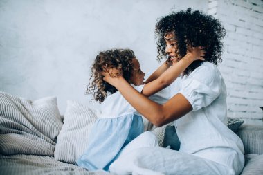 Beyaz elbiseli, kıvırcık saçlı bir anne, kıvırcık saçlı bir kız, saçlarına ve kafalarına dokunuyor. 