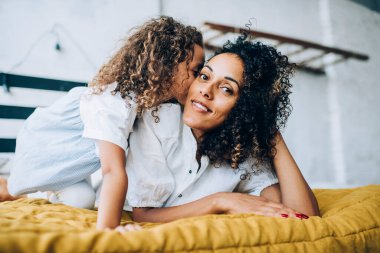 Yatakta dinlenip kameraya bakan kıvırcık saçlı, etnik pozitif bir kadının yanağına sarılıp öpen bir çocuk.