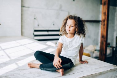 Şaşkın, sevimli, kıvırcık saçlı, günışığında tahta kürsüde bacak bacak üstüne atmış bir çocuk.