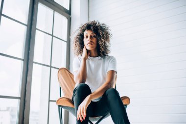 Duygusuz kıvırcık saçlı, günlük beyaz tişörtlü, mavi pantolonlu, açık renkli yaratıcı stüdyoda boyuna dokunan kadın.