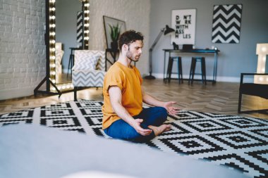 Turuncu tişörtlü ve mavi pantolonlu genç adam oturma odasında bacak bacak üstüne atmış Lotus pozisyonu üzerinde meditasyon yapıyor. 