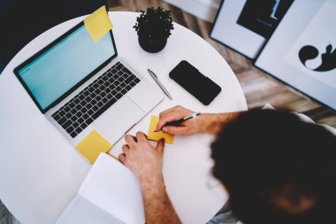 Ofisteki dizüstü bilgisayarlı cep telefonuyla masada oturan sarı etiketli kalemle notlar alan meçhul insanlardan.