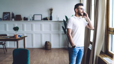 Koyu renk saçlı, beyaz tişörtlü ve kot pantolonlu genç adam telefonla konuşuyor ve geniş modern odada pencereden dışarı bakıyor.