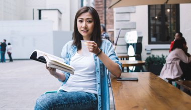 Esmer, güzel, Asyalı bir kadın soğuk, ferahlatıcı bir içecek içiyor ve açık hava kafesinde ahşap masadaki kameraya bakarken kitap okuyor.