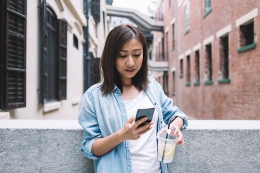 Uzun saçlı, kot ceketli, sosyal ağda mesajlaşan ve yürürken taş korkuluklara yaslanırken limonata içen Asyalı genç bayan.