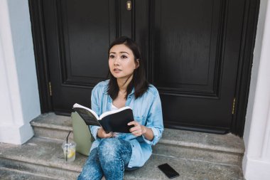 Kot pantolon giymiş, siyah kapılı merdivenlerde oturmuş kitap okurken şaşırmış bir şekilde yukarı bakan etnik bir kız öğrenci.