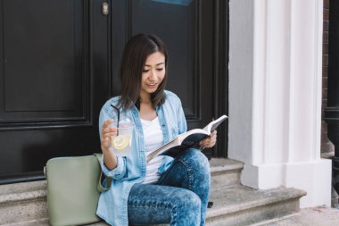 Kot pantolonlu mutlu Asyalı kız öğrenci elinde limonatayla merdivenlerde oturmuş kitap okurken.