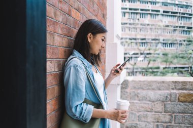 Tuğla duvara yaslanmış bir fincan kahve ve sosyal medyada sohbet eden konsantre ve odaklanmış bir etnik kadının yan görüntüsü