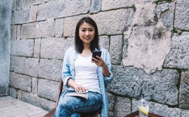 Havalı, neşeli, limonata bardağında limonatayla gezen, akıllı telefonuyla sokakta, tuğla duvarın yanında otururken mesaj atan genç Asyalı bir kadın.