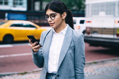 Resmi giyinmiş kendine güvenen girişimci şehir merkezinde duran akıllı telefondaki SMS mesajlarını daktilo ediyor. Moda gözlüklü kadın CEO 'su modern cep telefonuna 4G internet kullanarak uygulama yüklüyor.