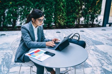 Resmi gözlüklü başarılı genç etnik bayan tablet üzerinde çalışıyor ve arka planda bulanık kafenin dışındaki masada oturuyor.