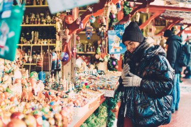Mutlu gülümseyen, iri yarı, siyah ceketli, çekici bir kadın. Noel festivali etrafında dolanıp renkli hediyeler seçiyor.
