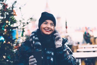 Güzel gülümseyen yeniyetme, kalın giysiler içinde genç bir kadın kış Noel fuarında kahve fincanıyla ayakta dururken akıllı telefonuyla konuşuyor.