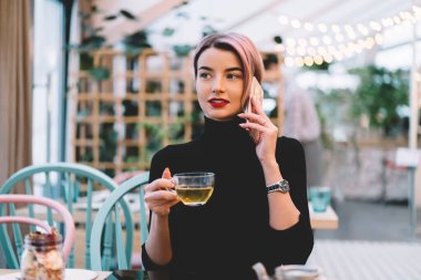 Havalı giyinmiş, pembe saçlı şık bir kadın akıllı telefondan konuşuyor ve hipster kafeteryasında oturmuş çay içiyor.