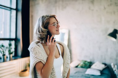 Klasik gözlüklü çekici bir kadın cep telefonu uygulaması ile en iyi arkadaşınla iletişim kurmaktan zevk alıyor. Gözlüklü, gülümseyen bir kadın evde cep telefonuyla konuşuyor.