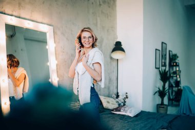 Ayna yanında gözlüklü mutlu genç bayanın yan görüntüsü ve kameraya bakarken telefonla konuşan geniş yatak.