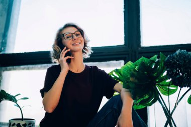 Optik gözlüklü mutlu kadın portresi ev penceresinin yanında oturuyor ve boş zamanlarında kameraya gülümsüyor cep telefonu cihazı ile en iyi arkadaşla iletişim kurmak için, uygulamayla telefon eden neşeli kadın.