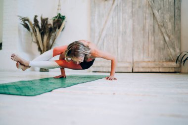 Güzel, neşeli atletik kadın spor kıyafetleri içinde, ahşap zeminli ve yeşillikli, parlak dekore edilmiş çatı katında, minder üzerinde meditasyon yapıyor.